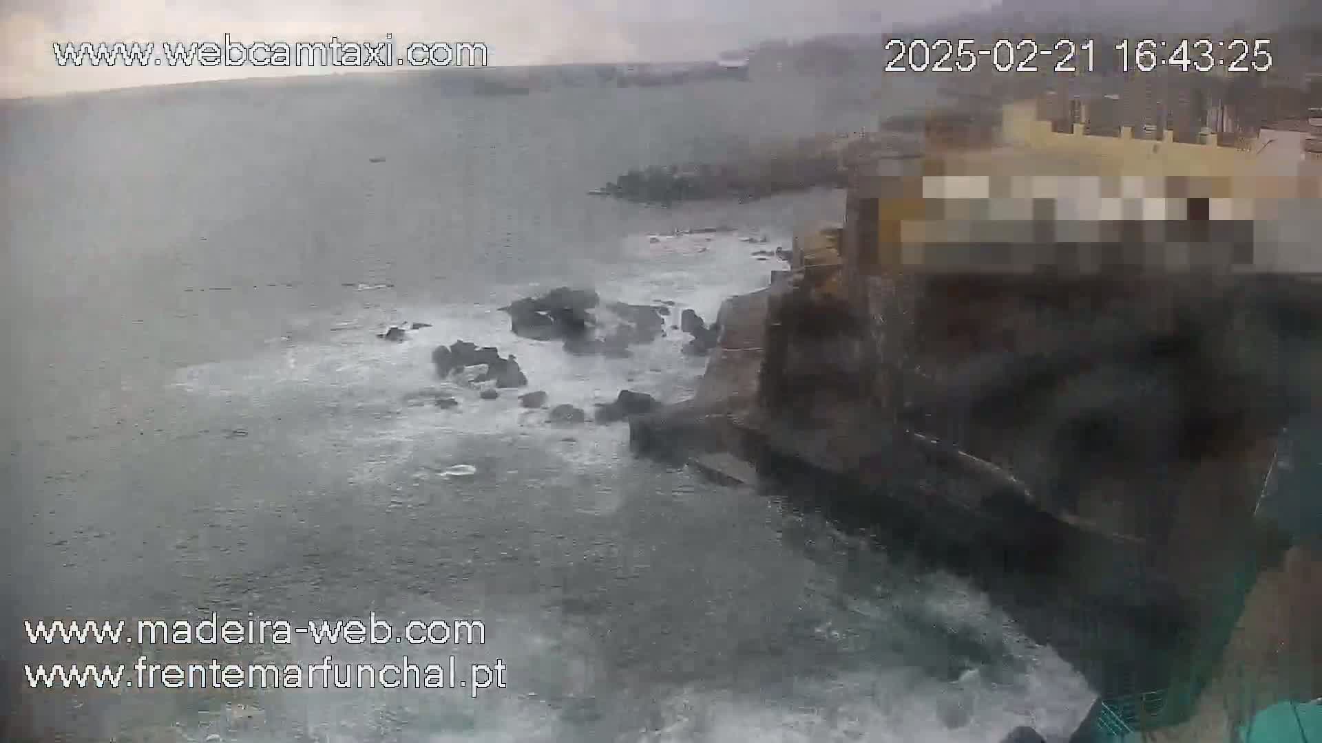 Waves crash against dark rocks near a light-colored building in stormy weather.