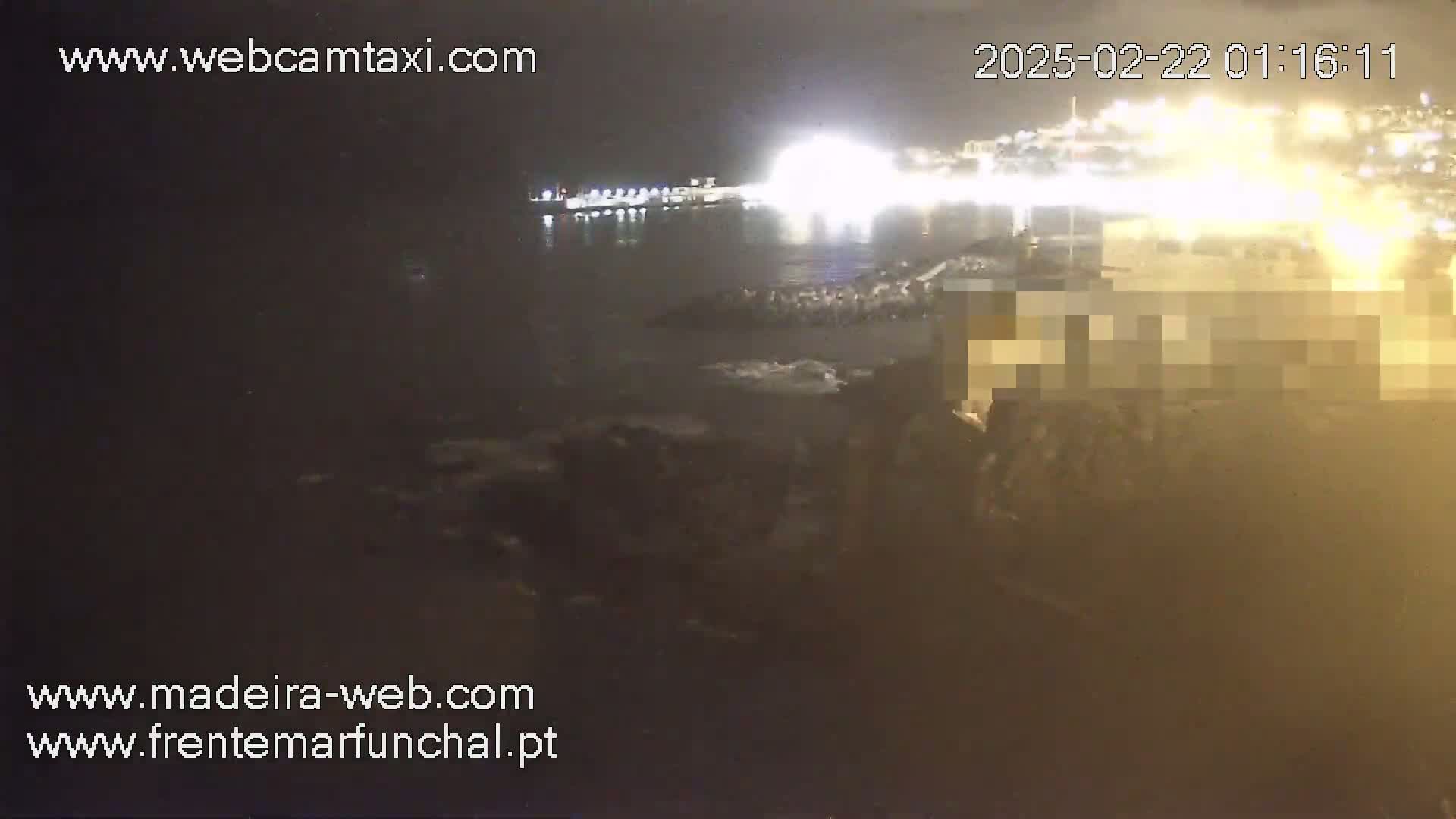 A nighttime, low-light view of a harbor with brightly lit buildings across the water, and dark, rocky shoreline in the foreground.