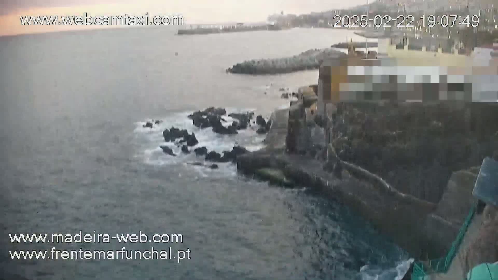 Ocean waves crash against dark rocks near a building on a cloudy day.