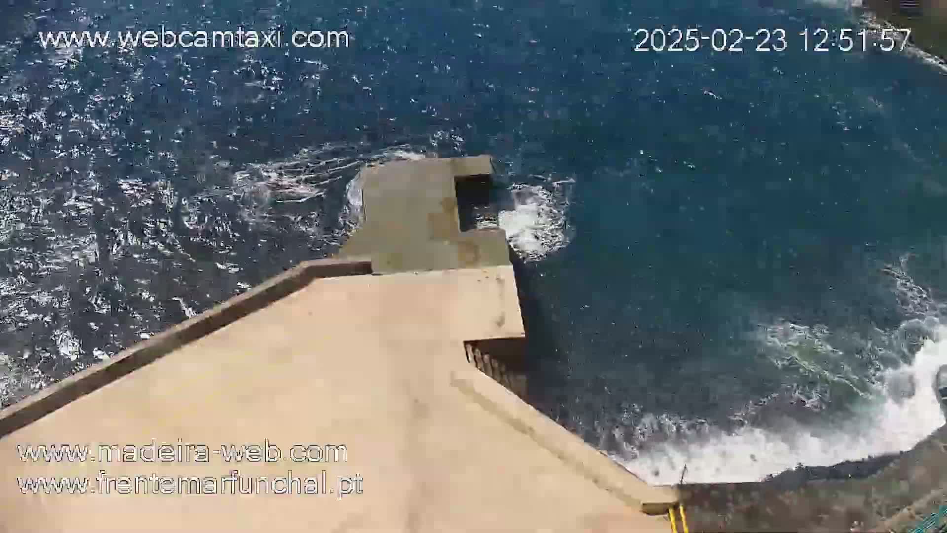 An aerial view shows a concrete structure extending into a dark blue ocean with moderate waves crashing against its edges.
