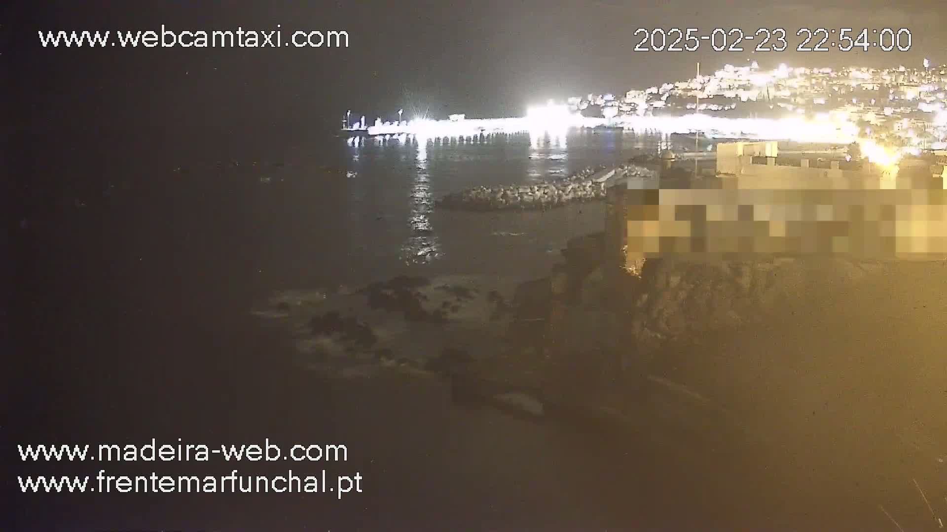 A nighttime view of a brightly lit waterfront area with dark, wavy water lapping against rocks.