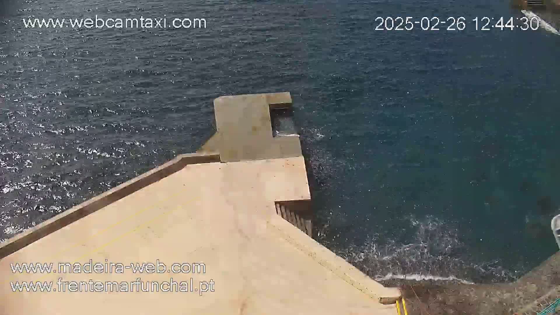 A tan concrete structure extends over dark-blue water with small waves splashing against it.