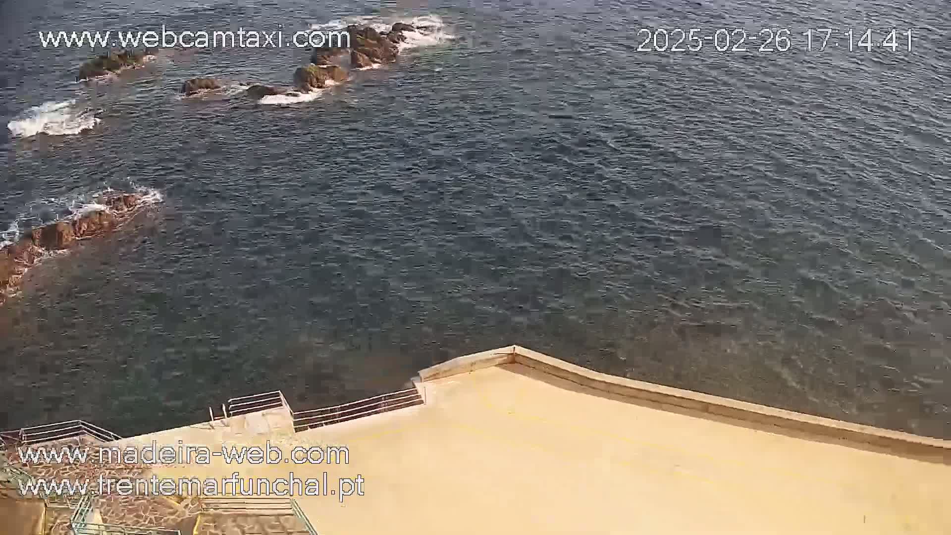 A tan-colored concrete platform overlooks dark-blue ocean water with small rocky outcrops and gentle waves.
