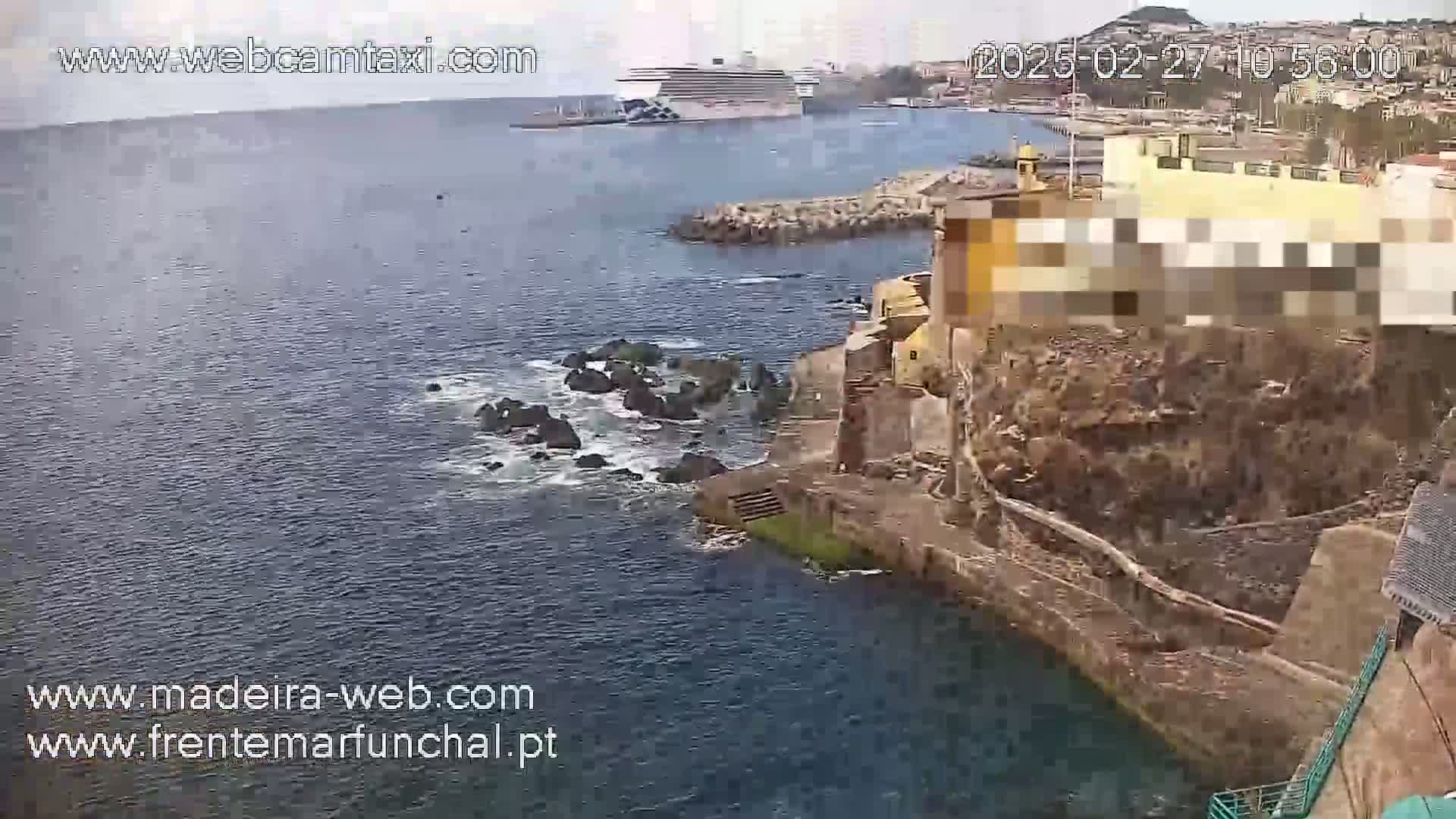 A partly cloudy day shows a rocky coastline with a building and a cruise ship in the distance, with waves breaking on the rocks.