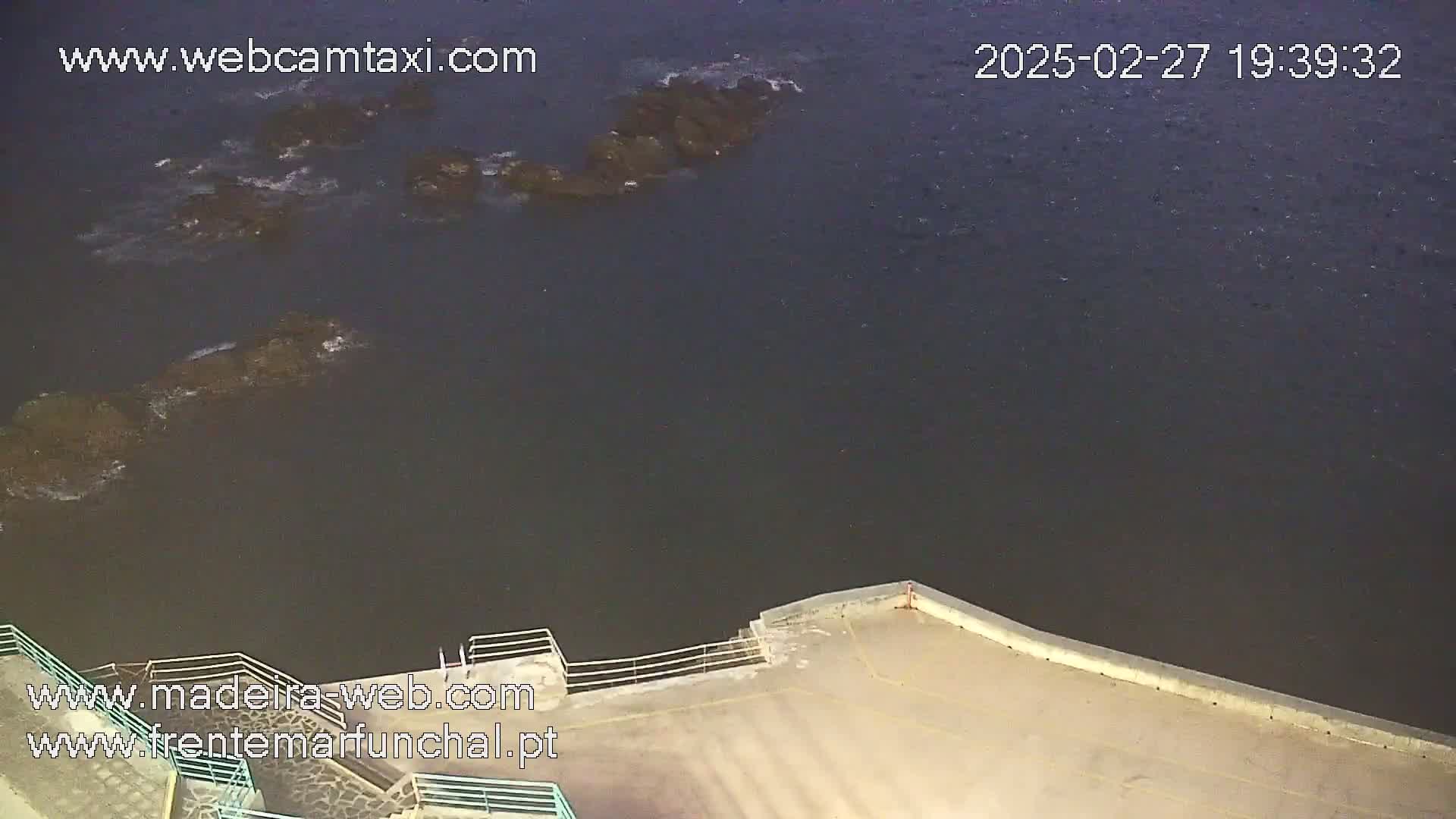 A dark body of water at night with several dark rocks partially submerged near a cement walkway with a railing.