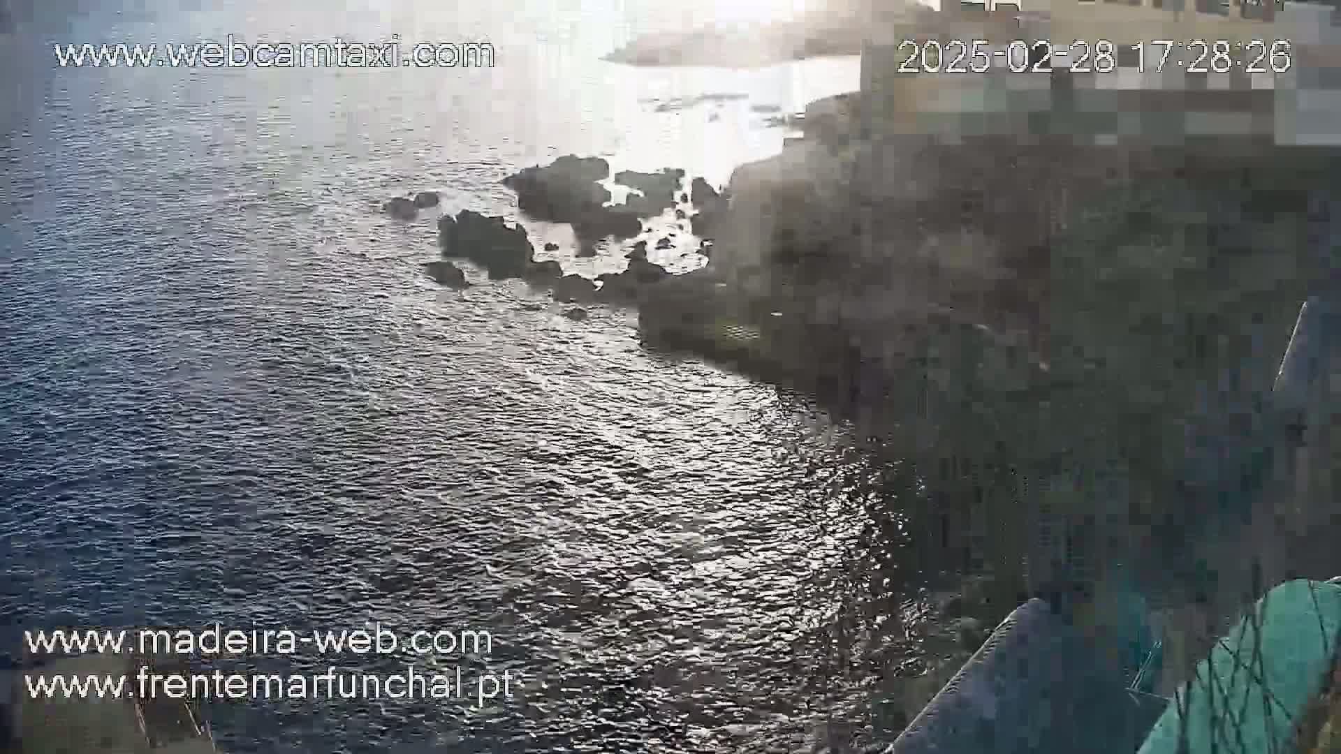 A rocky shoreline meets a dark, rippling ocean under a bright, possibly sunny sky.