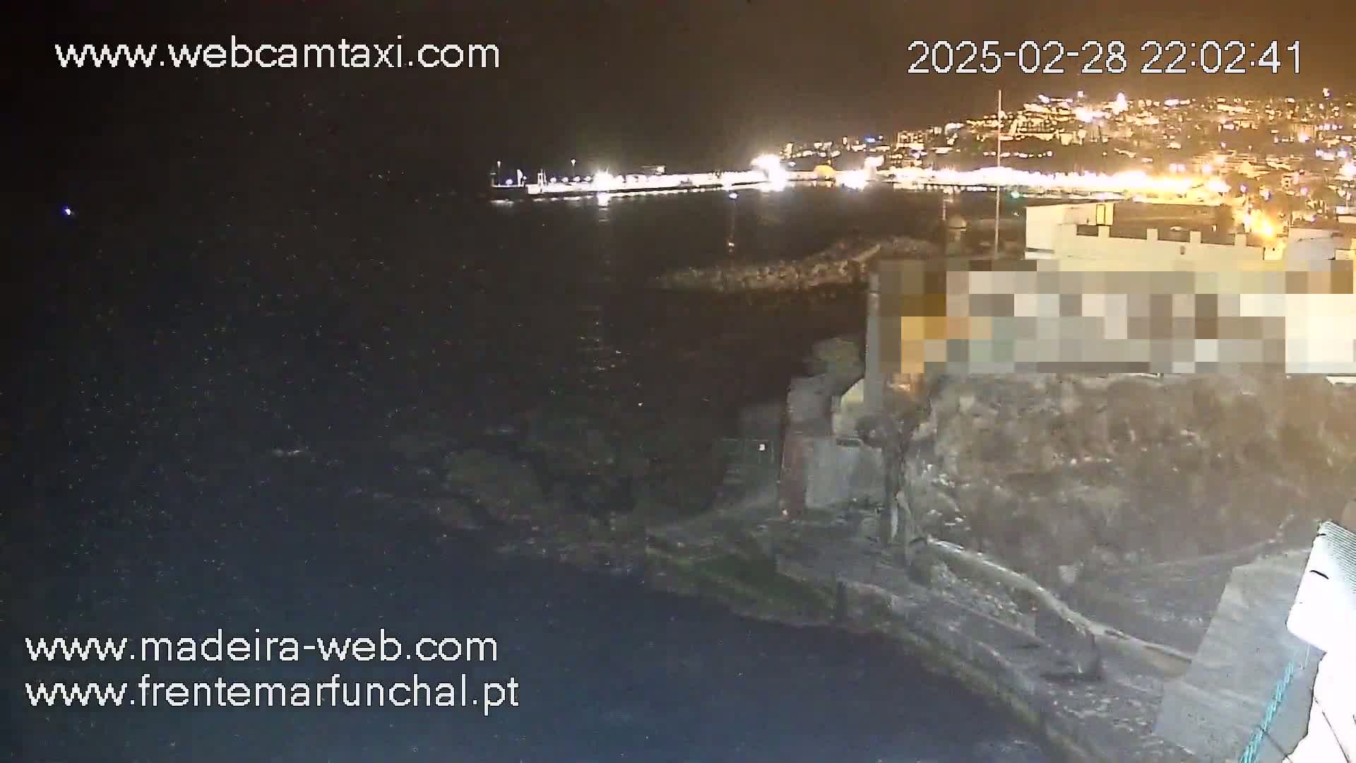 A nighttime view of a dark, rocky coastline with a brightly lit city in the distance across the water.