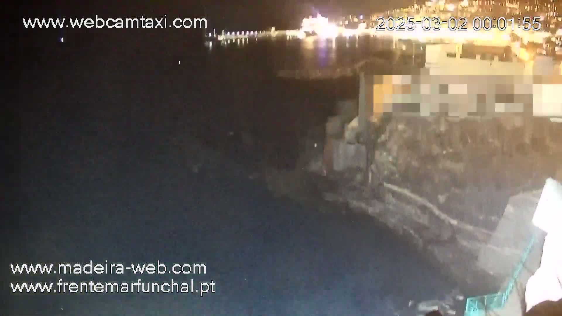 A nighttime, low-angle view shows a dark body of water next to a rocky shore with a building partially obscured, under a clear, dark sky with distant lights.