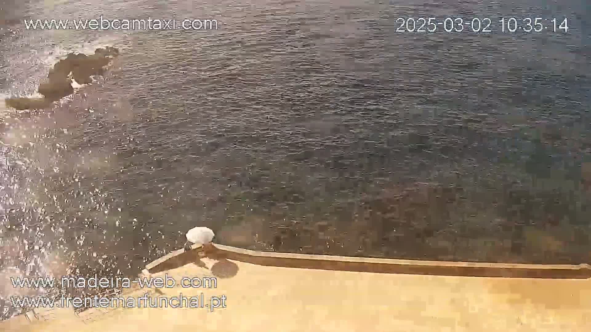 Waves are splashing against a low tan wall next to dark-colored water.