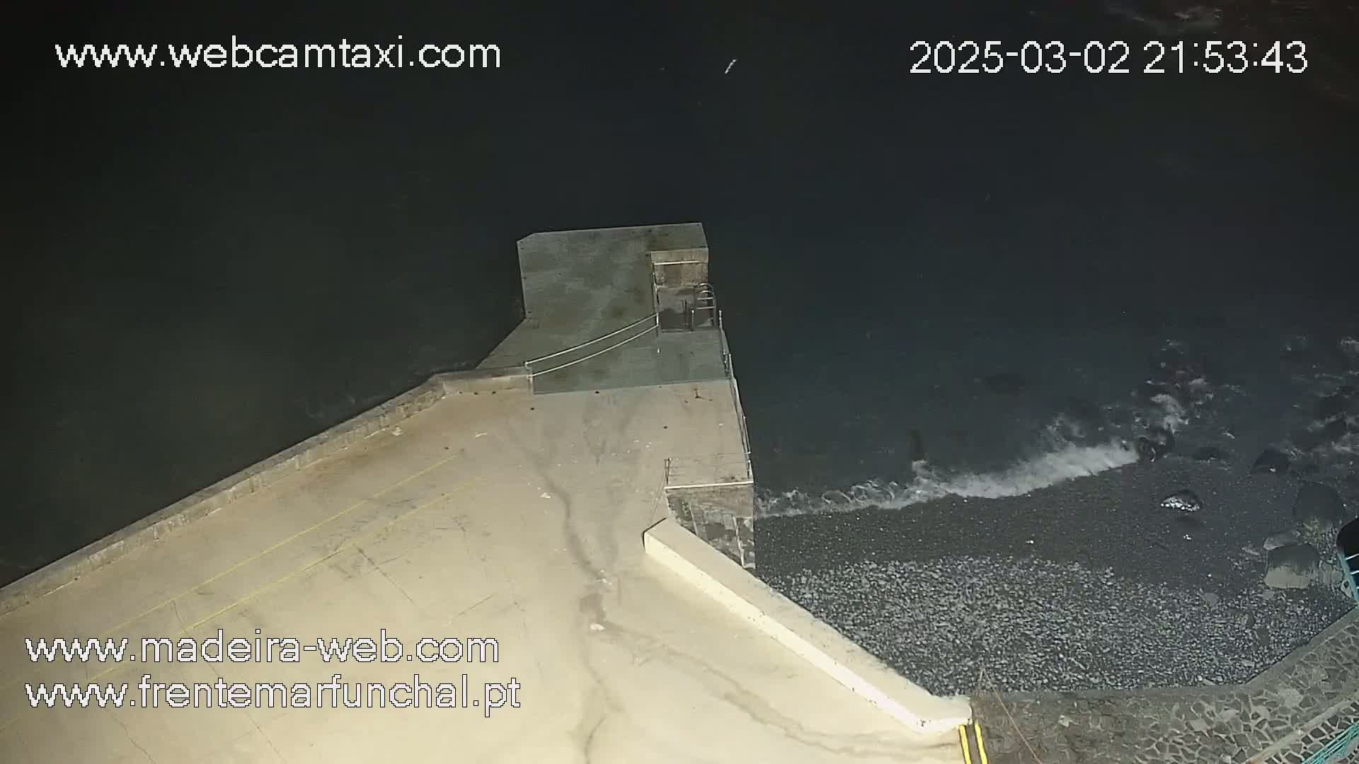 A nighttime view of a concrete structure extending into dark, wavy water lapping against a pebble shore.