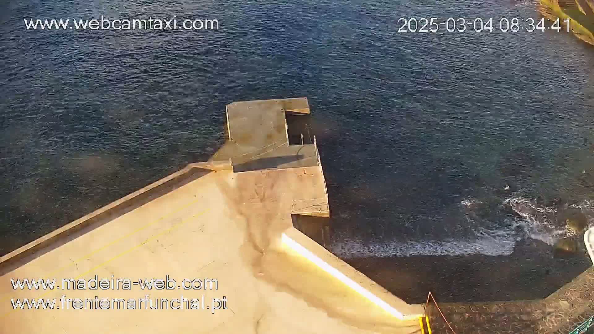 An aerial view shows a concrete pier extending into dark-blue water with small waves lapping against it on a sunny day.