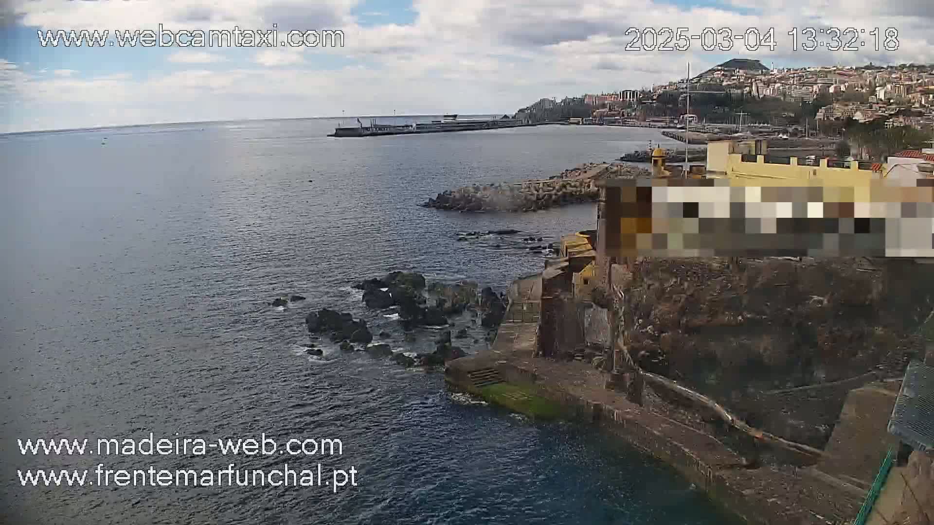 A partly cloudy day overlooking a rocky coastline and calm ocean, with a town visible in the distance.