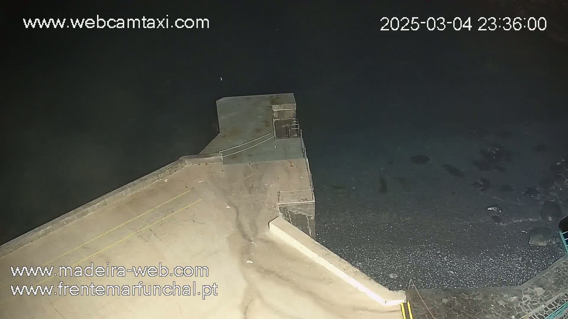 A nighttime, high-angle view shows a concrete structure extending into dark water, with a rocky shoreline visible below.