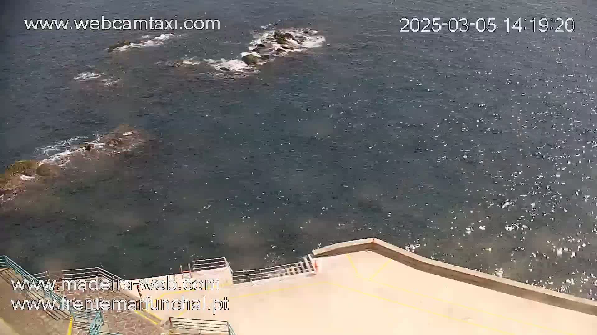 An aerial view shows dark-blue water with small waves lapping against a tan concrete structure with stairs leading down to the water's edge, and dark rocks visible just offshore.
