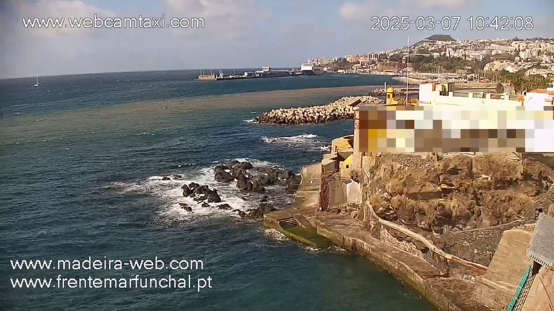 A coastal scene on a sunny day shows dark-blue water lapping against rocky shores and a building on a cliff overlooking a harbor with boats and a city in the distance.