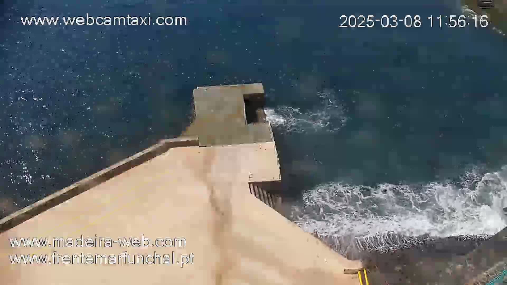 A concrete structure extends from a tan-colored area into dark-blue water with white-capped waves crashing against its base.