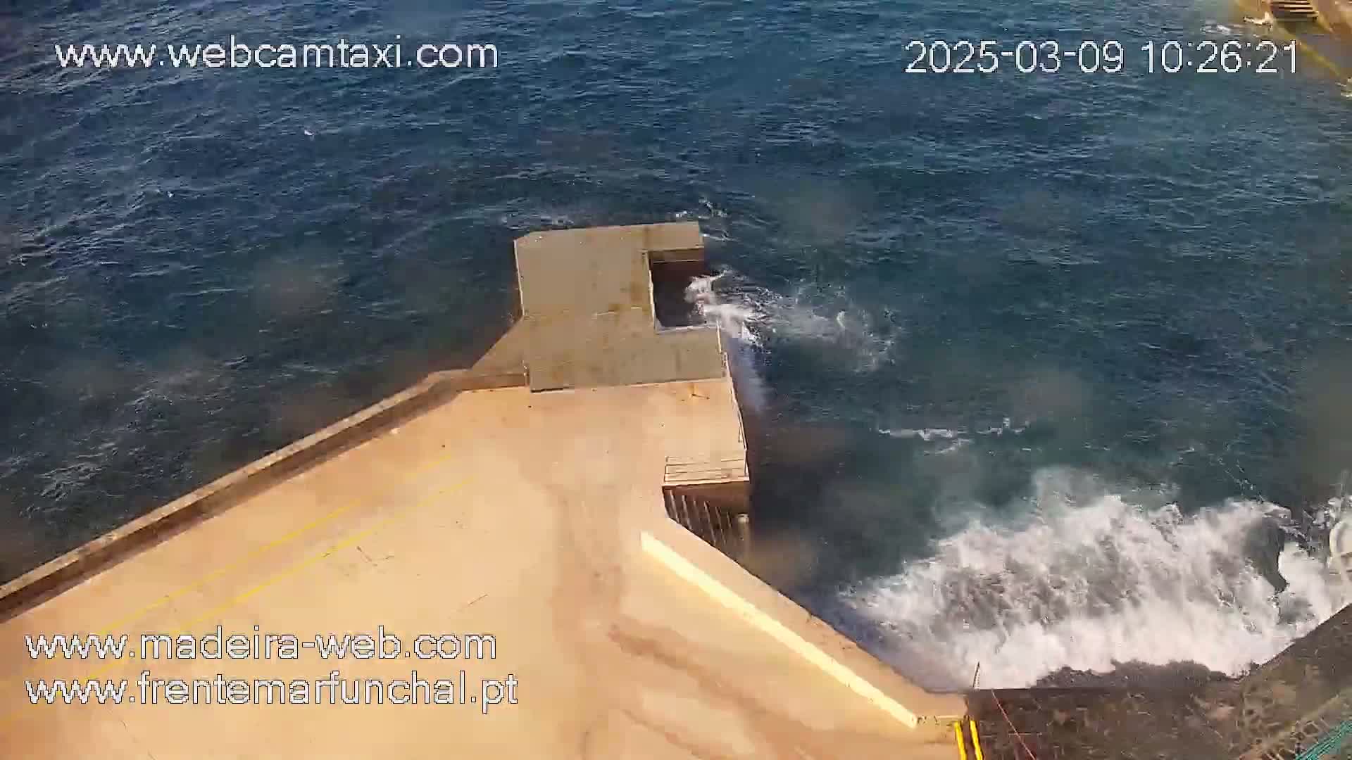 An overhead view shows a concrete structure extending into a dark blue ocean with moderately strong waves crashing against its edge.