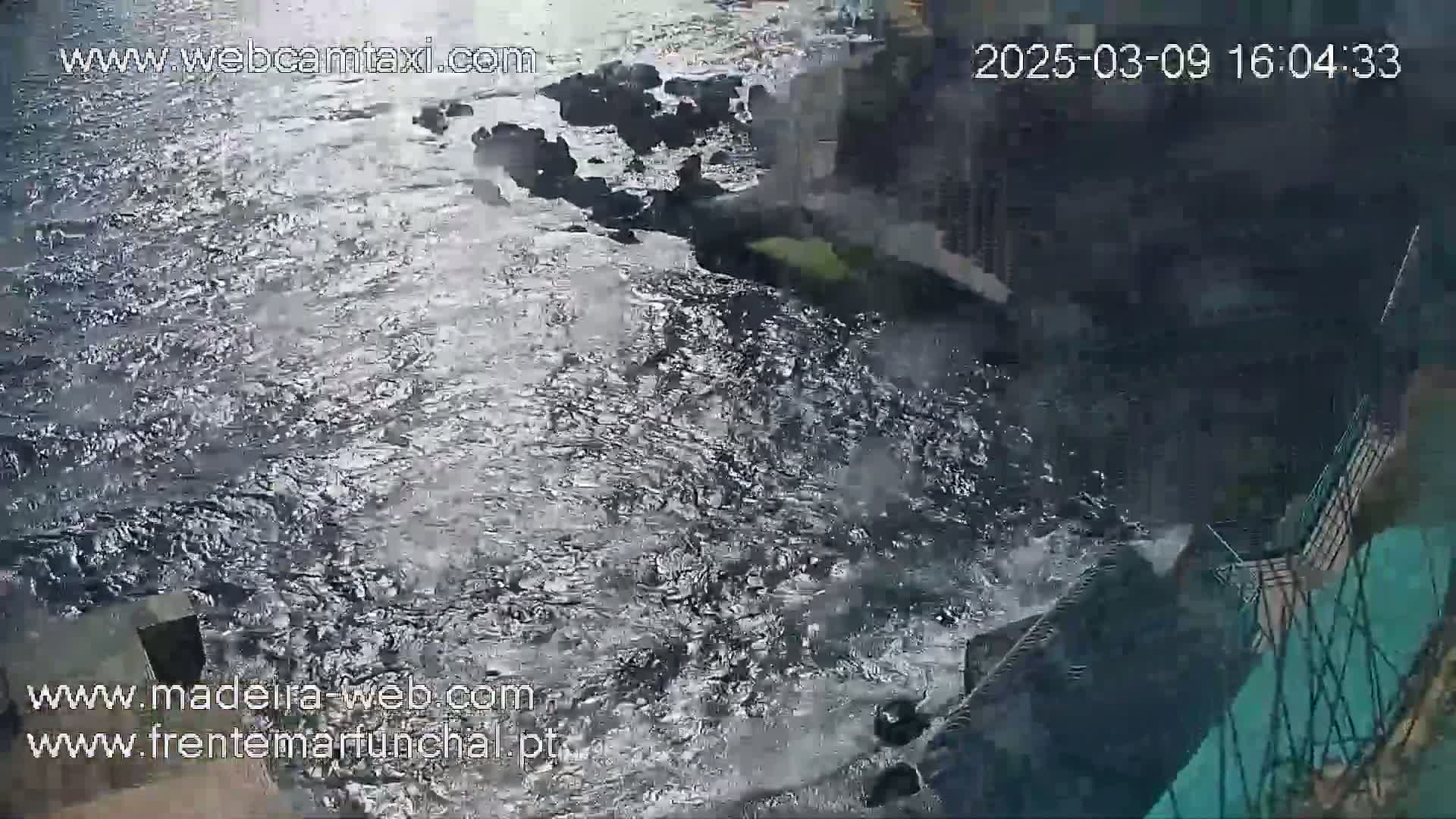 Turbulent dark water flows over dark rocks near a dark structure, possibly a wall or embankment.