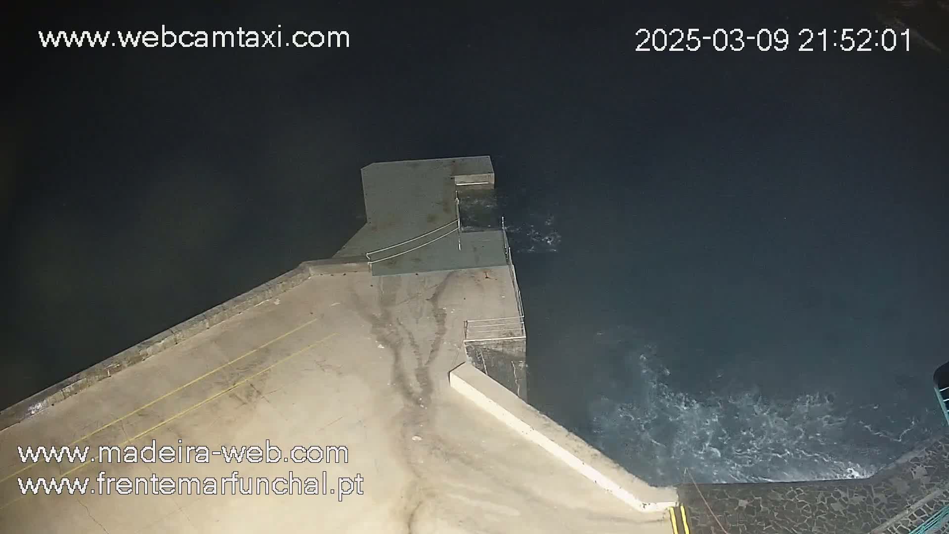 An aerial nighttime view shows a concrete platform extending into dark water with small waves crashing against it.