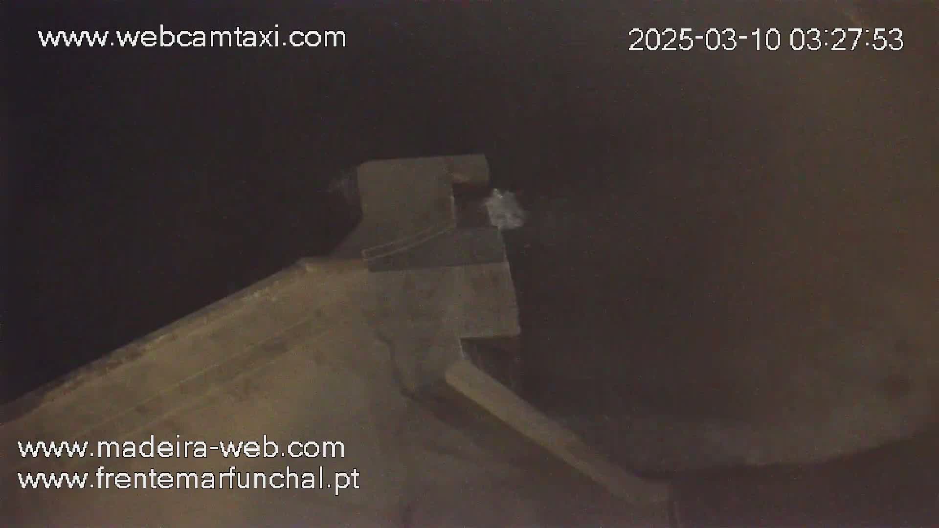 A nighttime, low-angle view shows a section of a dark gray concrete structure, possibly a wall or pier, with faint white splashes possibly from water visible in the background.