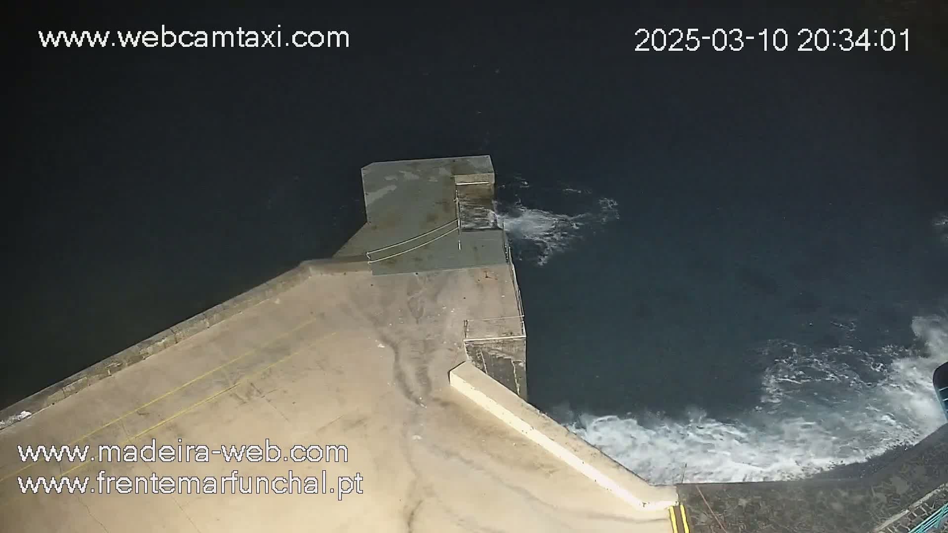 A nighttime, high-angle view shows waves crashing against a concrete seawall.