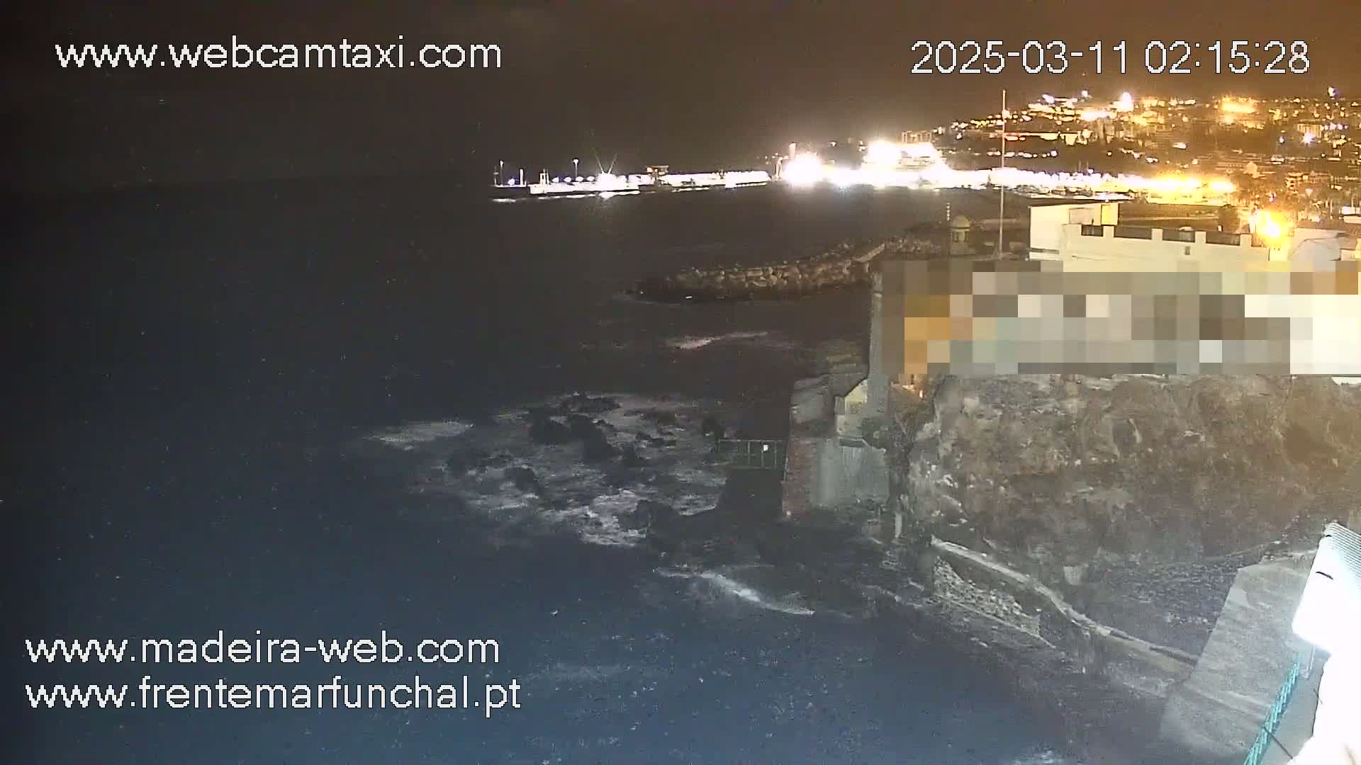A nighttime view of a rocky coastline with dark, slightly choppy water, illuminated by lights from a city in the distance.