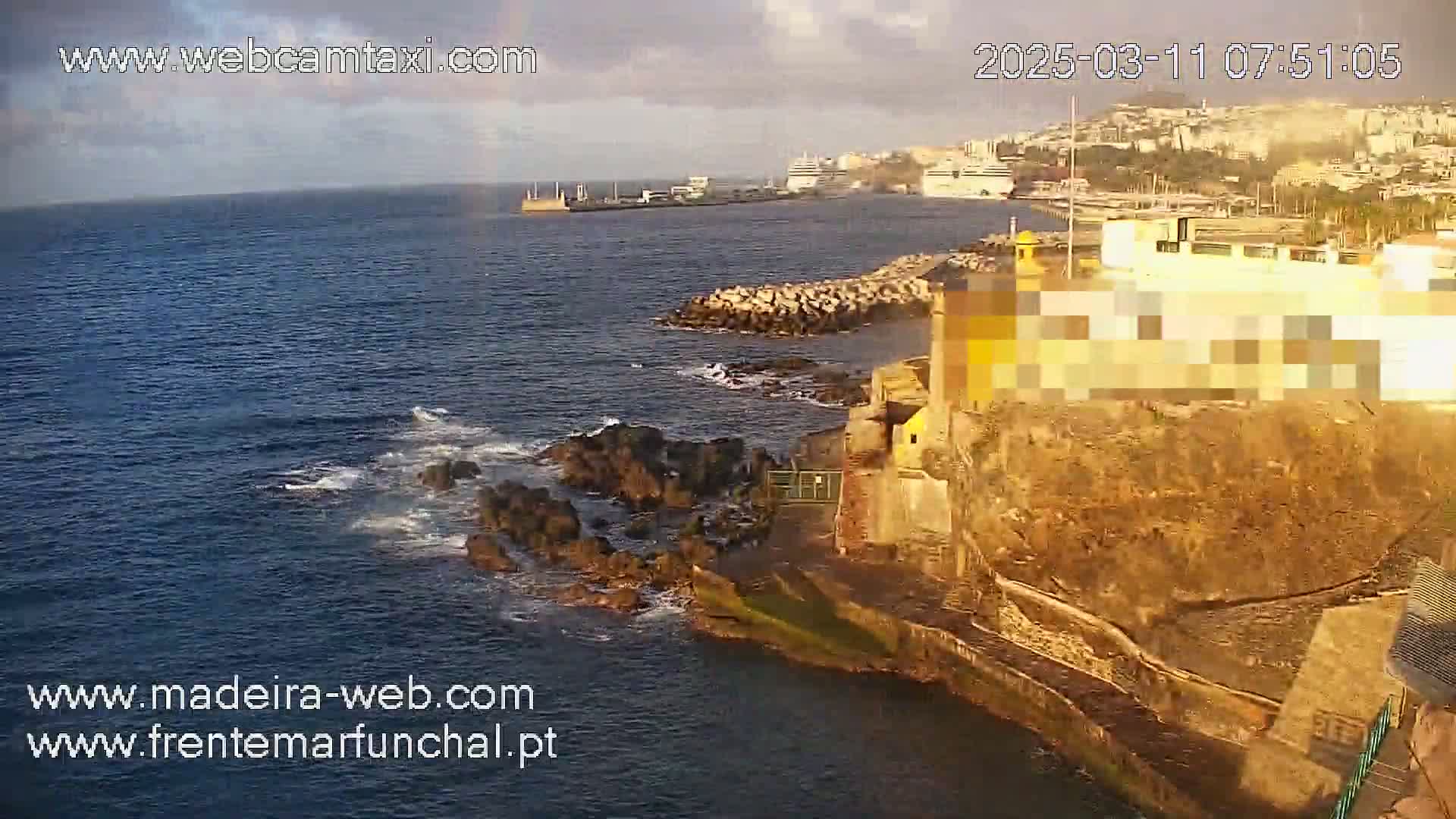 A rocky coastline with a building and a breakwater is shown, with calm ocean waves and a partly cloudy sky.