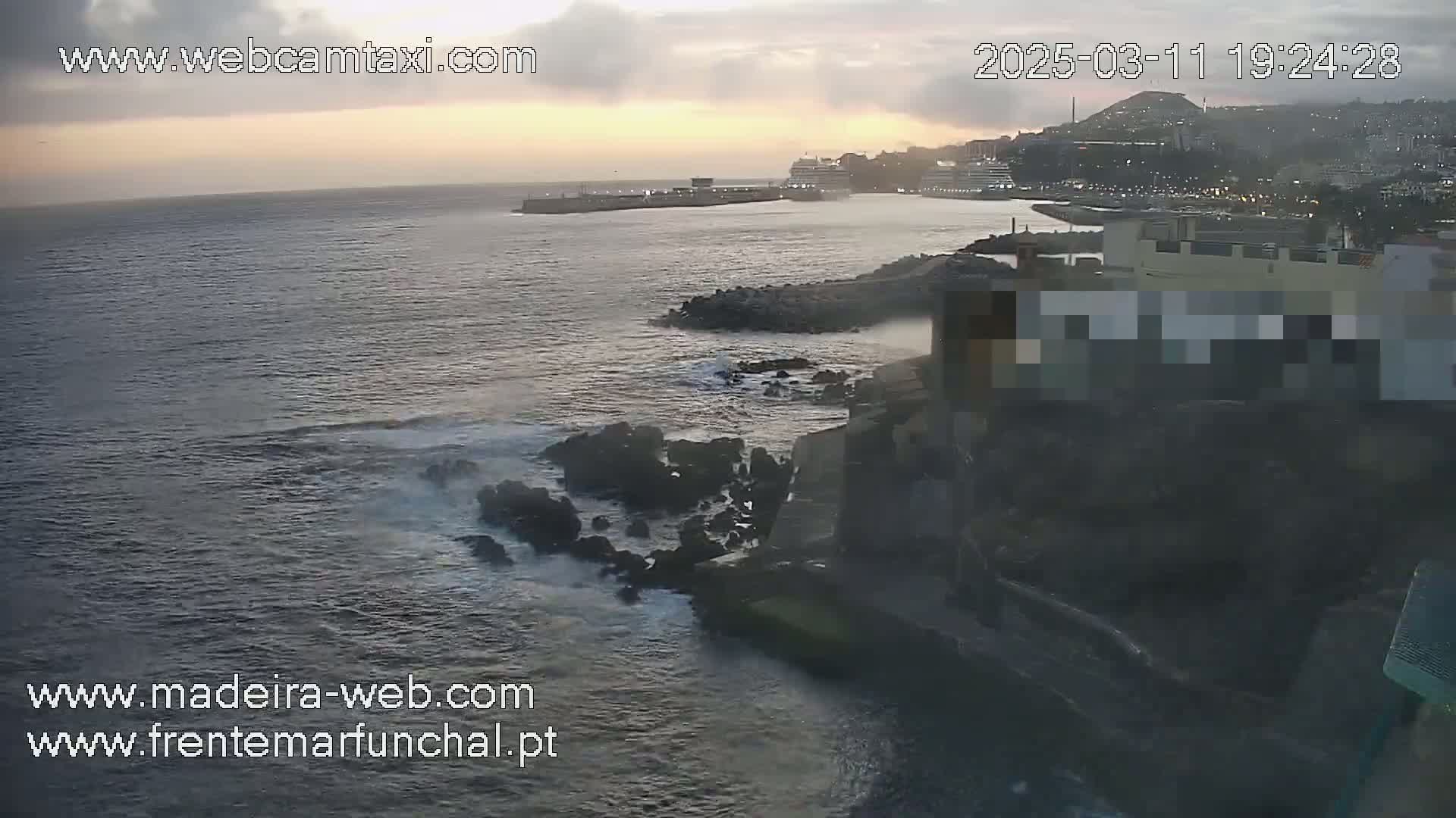 A coastal scene at sunset shows dark, rocky shores meeting a dark ocean with small waves, and a city in the distance under a cloudy sky.