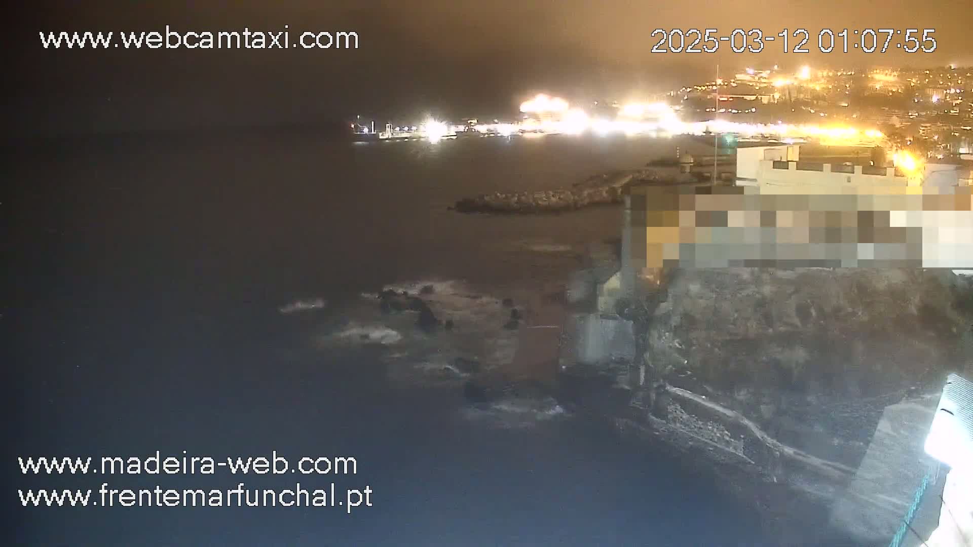 A nighttime coastal scene shows a dark body of water with waves lapping against a rocky shore, near a brightly lit city in the distance.