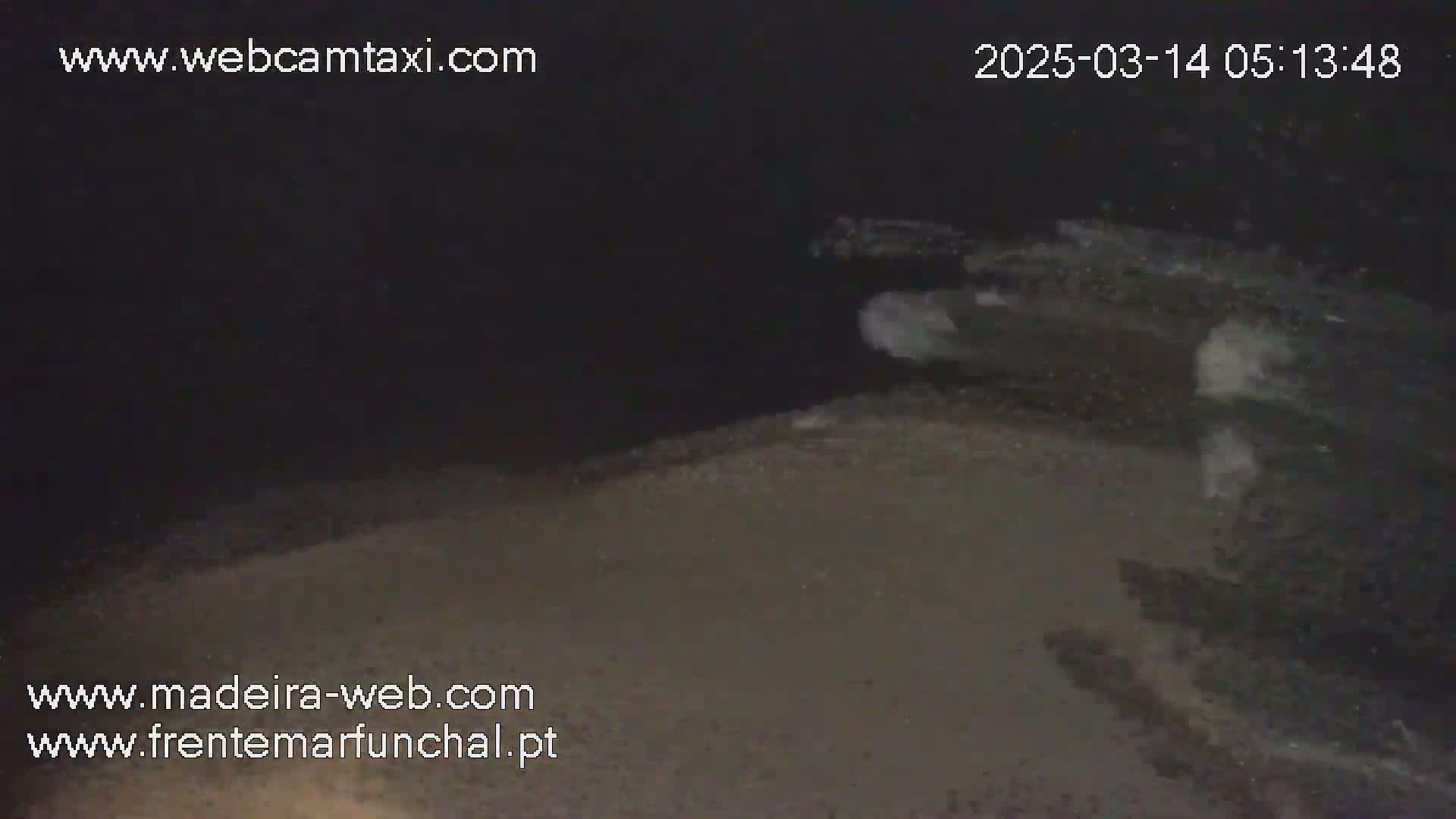 A nighttime, low-light view of a dark, sandy or gravelly surface with some lighter patches that may be water or foam.