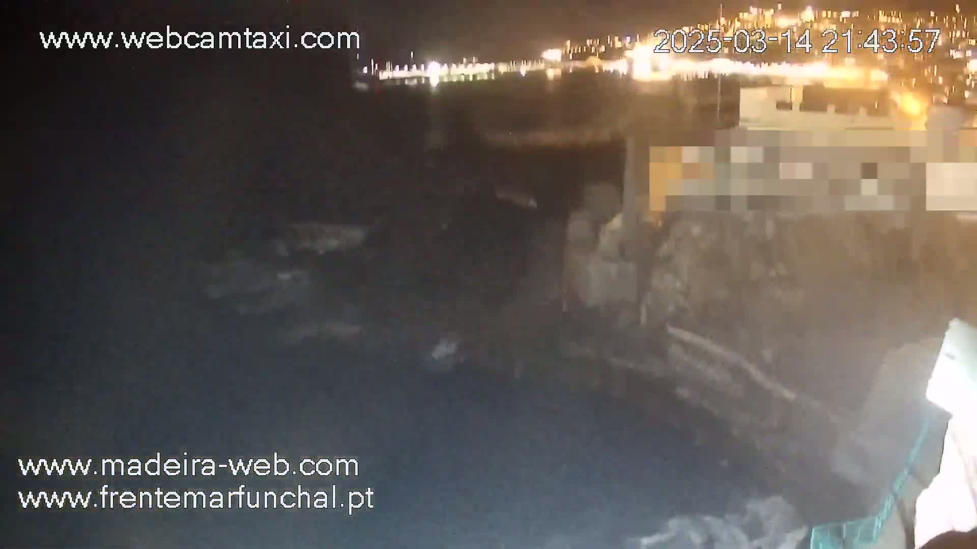 A nighttime, low-light view of a dark, rocky shoreline with a blurred city skyline and lights in the distance.