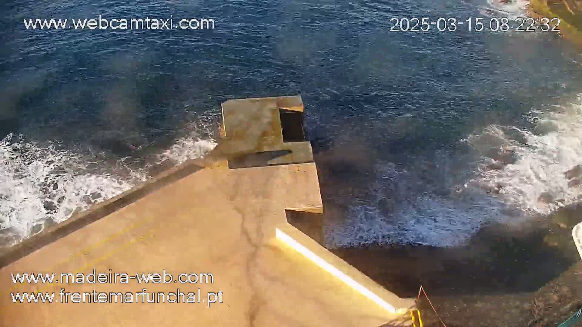 A high-angle view shows a concrete structure extending over dark-blue water with white-capped waves crashing against it.