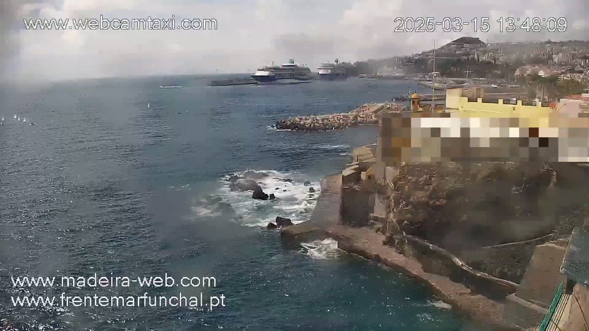 Two cruise ships are in a harbor on a partly cloudy day, near a rocky coastline with a building, and some small boats are visible in the water.