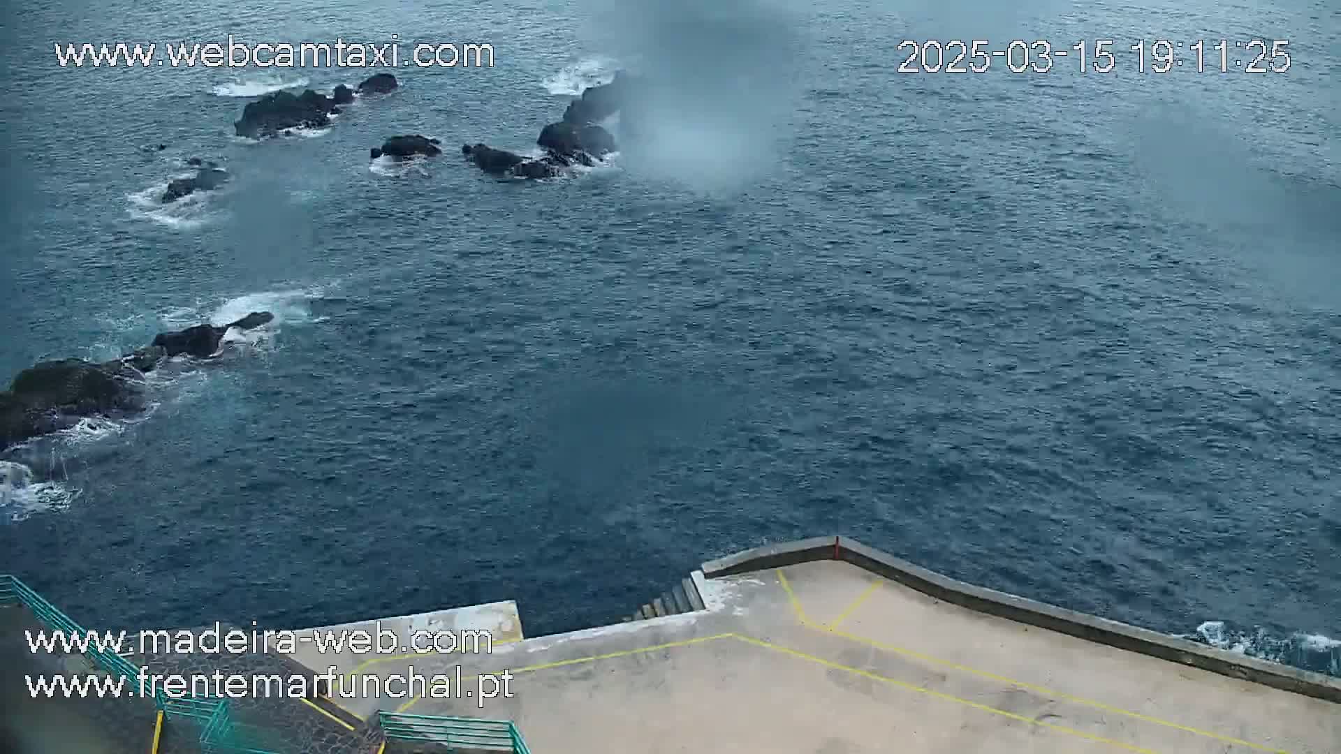Dark-blue ocean water laps against dark rocks near a concrete platform on an overcast day.