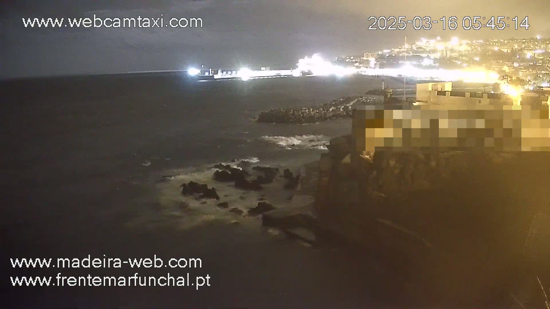 A large ship is docked at a nighttime harbor, with slightly choppy water visible near a rocky shoreline and a city with lights in the background under a dark, cloudy sky.