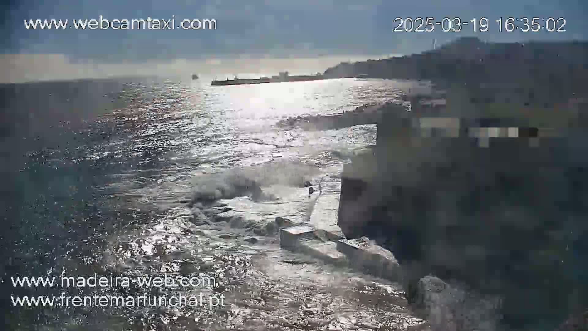Waves crash against a rocky shoreline under a partly sunny sky.