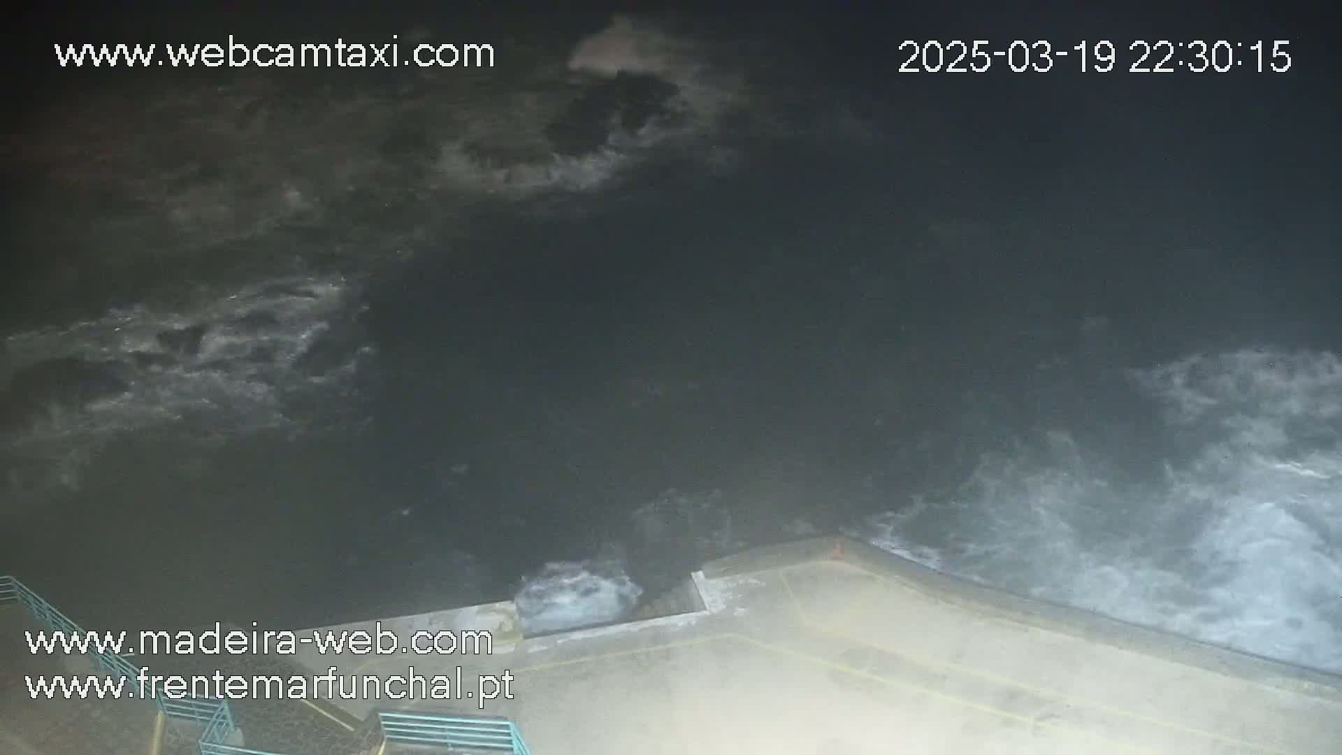A nighttime view of dark, choppy water crashing against a concrete wall under a cloudy sky.
