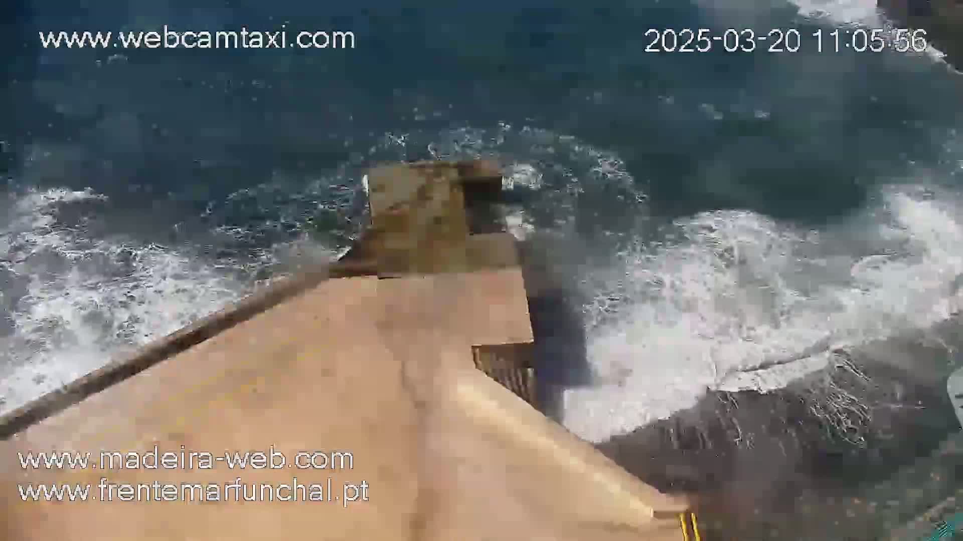 Ocean waves crash against a tan-colored concrete structure near a rocky shoreline.