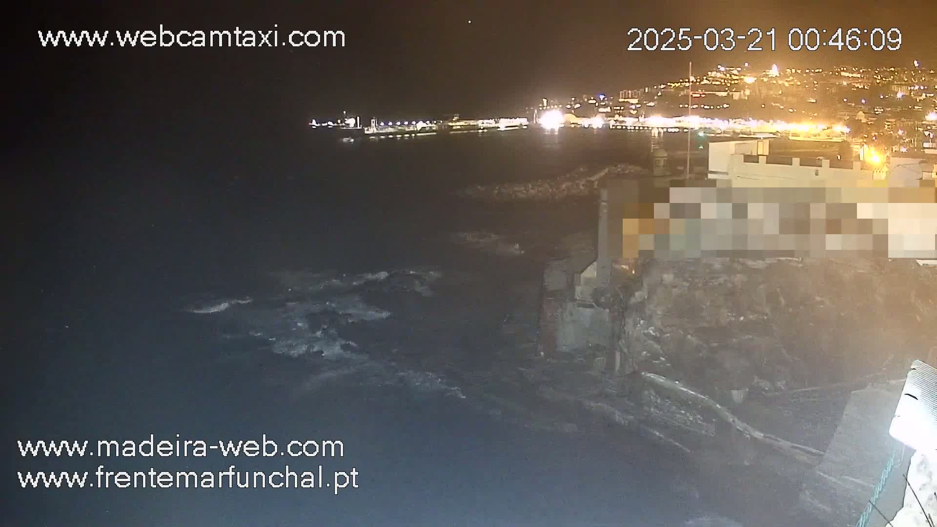 A nighttime coastal scene shows dark, choppy water lapping against a rocky shore, with a brightly lit city visible in the distance.