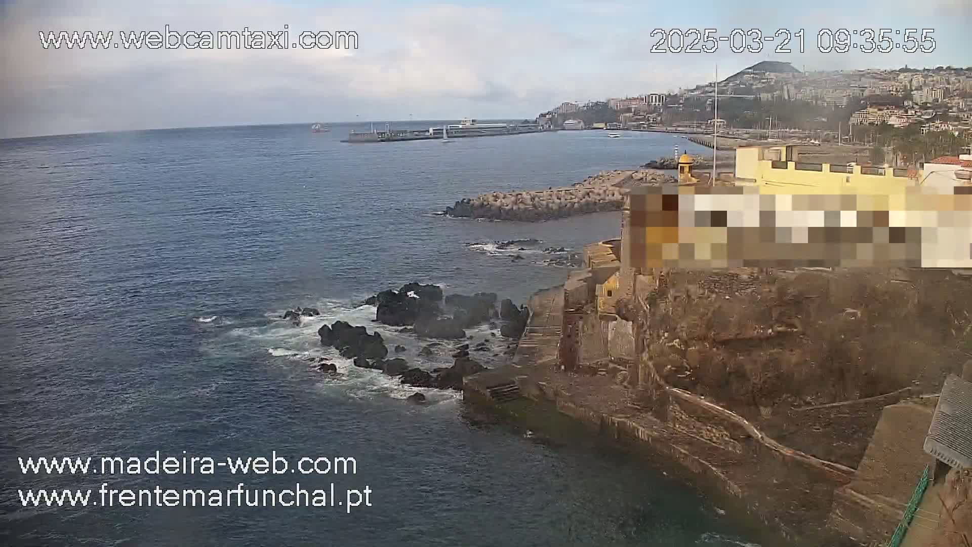 An overcast day shows a rocky coastline with a partially-visible yellow building, a body of water with some small waves, and a city visible in the distance.