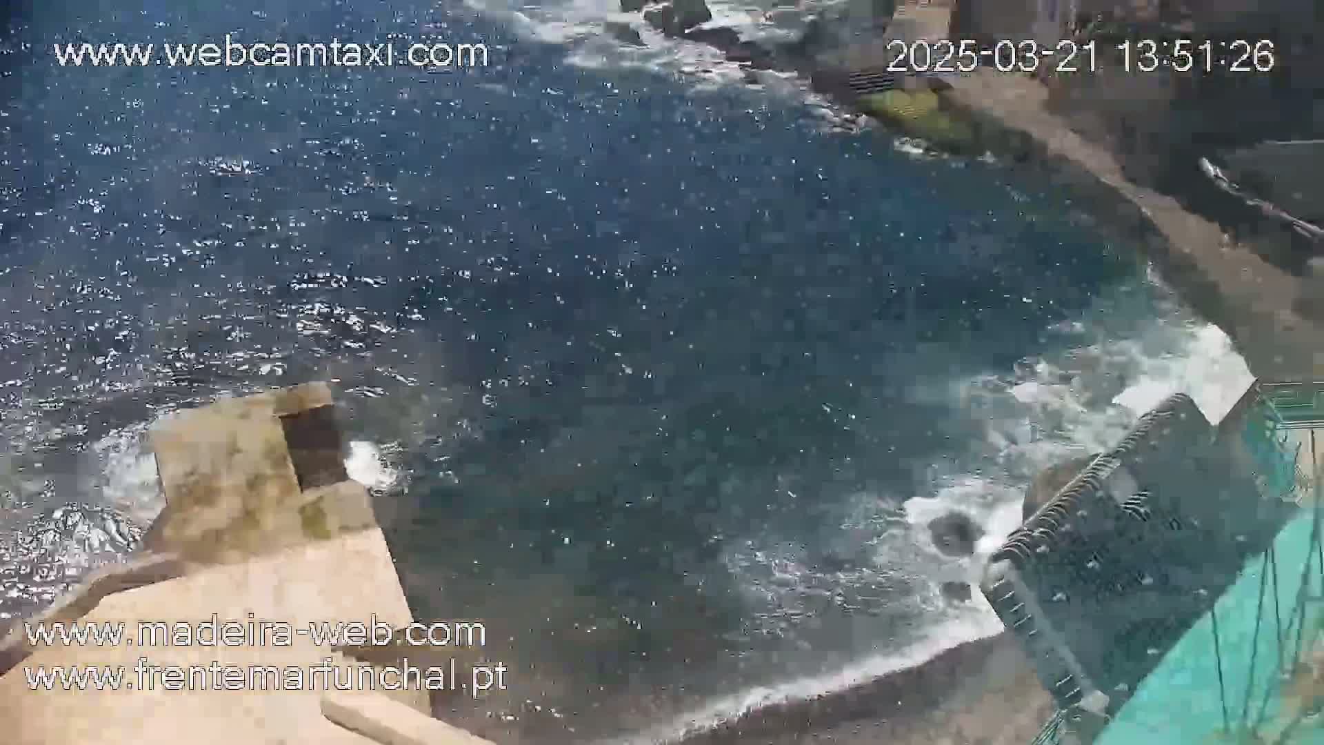 Dark-blue water crashes against a light-brown concrete structure near a rocky shoreline.
