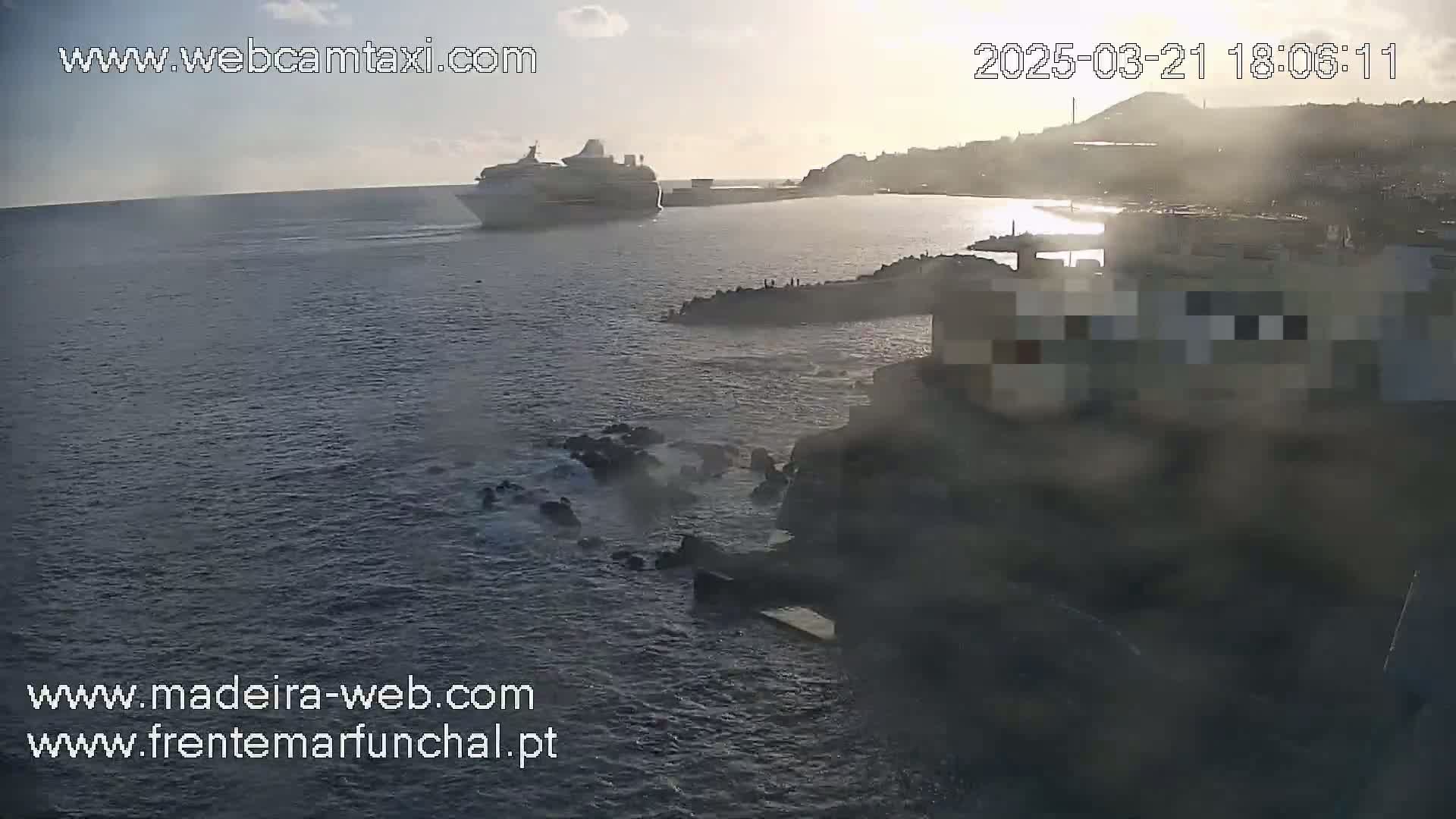 A large cruise ship is visible on calm water near a rocky coastline at sunset, with a hazy or foggy atmosphere.
