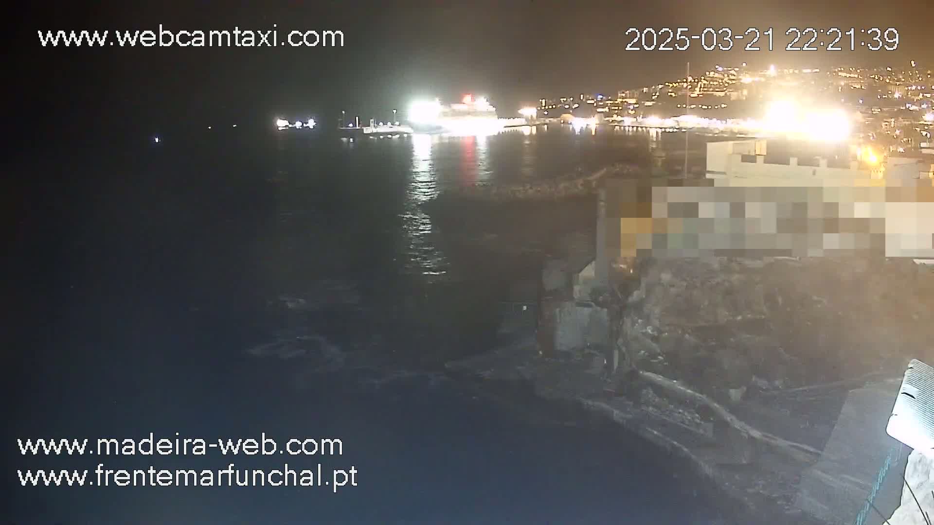 A nighttime view of a harbor with a large ship docked, illuminated buildings across the water, and dark, slightly choppy water.