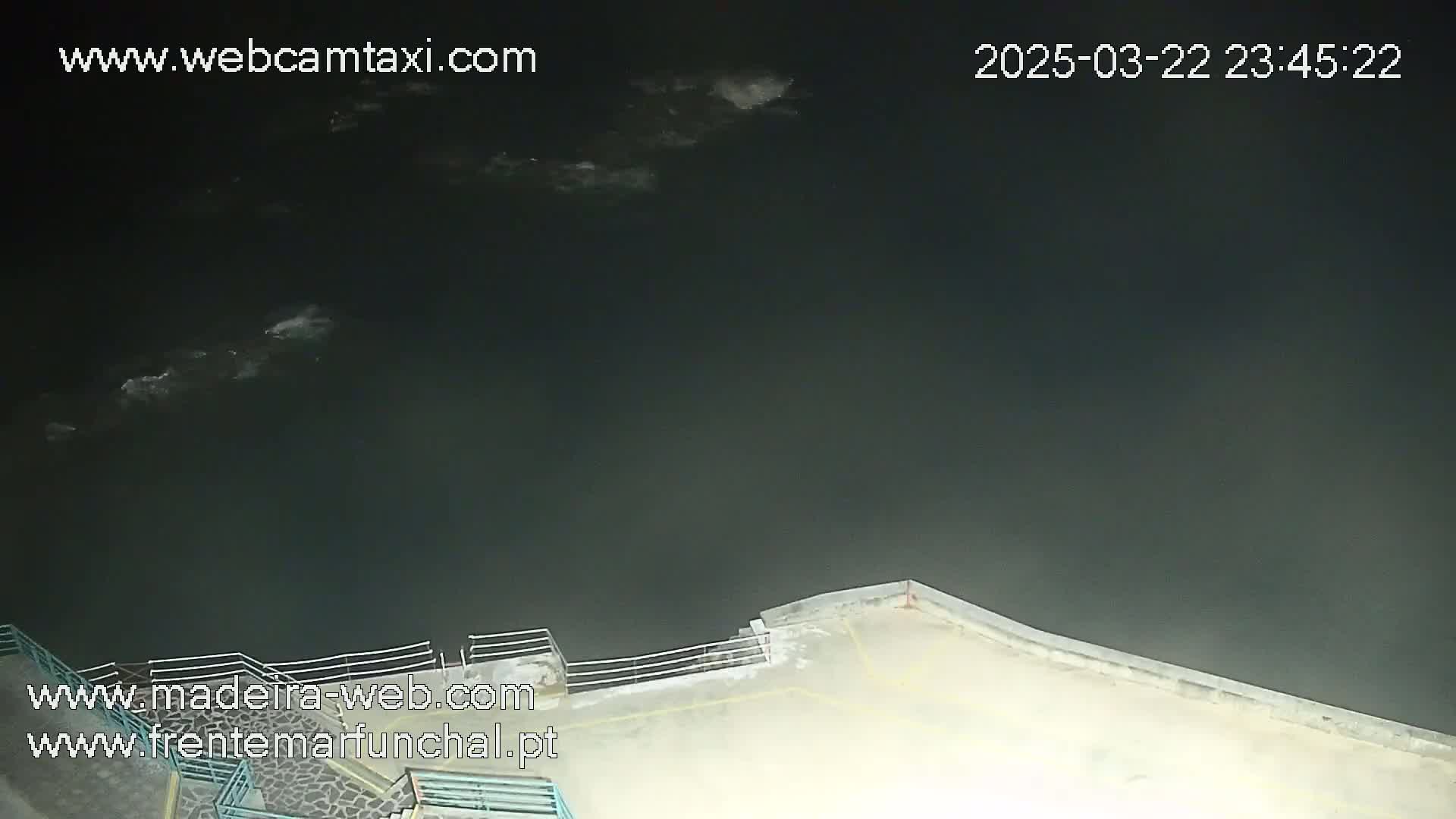 A nighttime, high-angle view shows a dark body of water meeting a paved area with a railing, under a mostly cloudy sky.