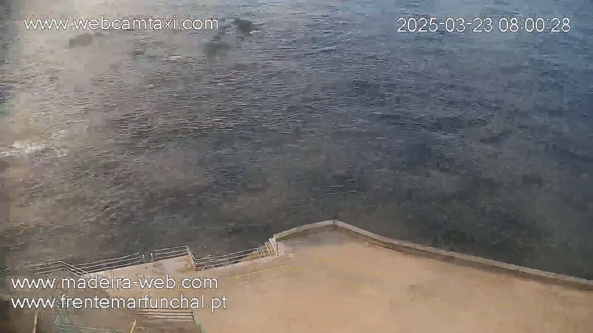 A high-angle view shows a dark-grey body of water meeting a tan concrete area with stairs leading down to the water.