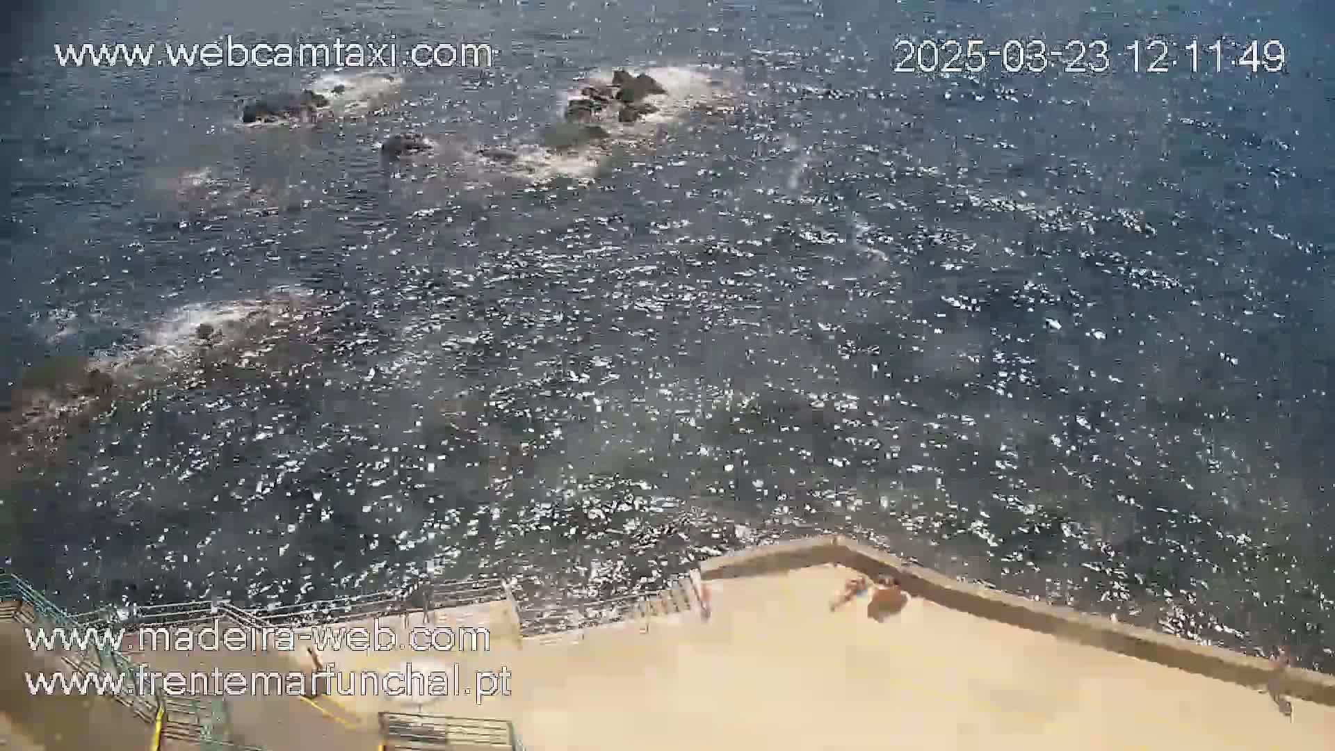 A high-angle view shows a rocky shoreline with choppy water and a few people sunbathing on a concrete platform.