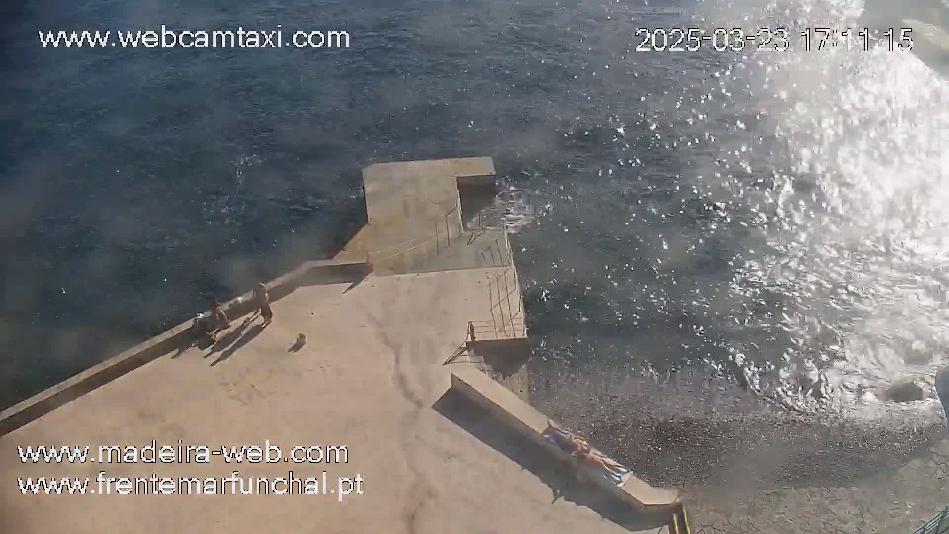 A concrete platform near a rocky shore shows several people relaxing and a small dog, with waves splashing against the platform on a sunny day.