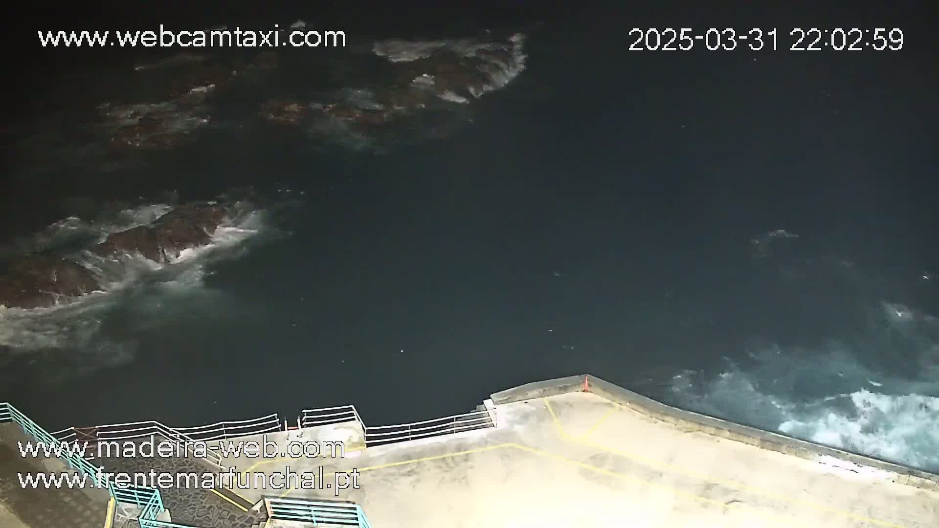 A nighttime, high-angle view shows dark ocean waves crashing against dark rocks near a concrete structure with a railing.