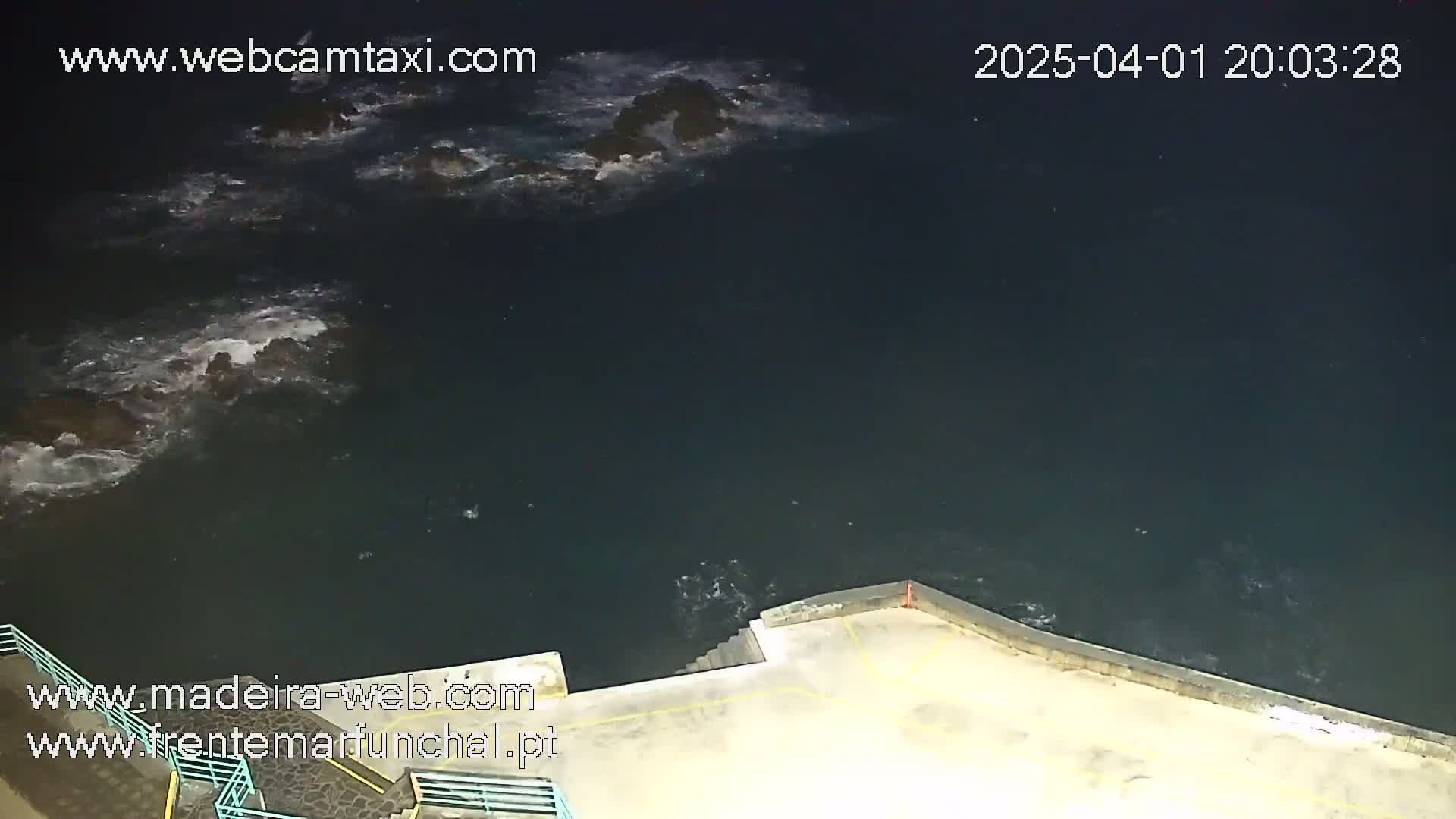 A nighttime view of dark water with small waves lapping against a concrete structure, featuring rocks partially submerged in the water.