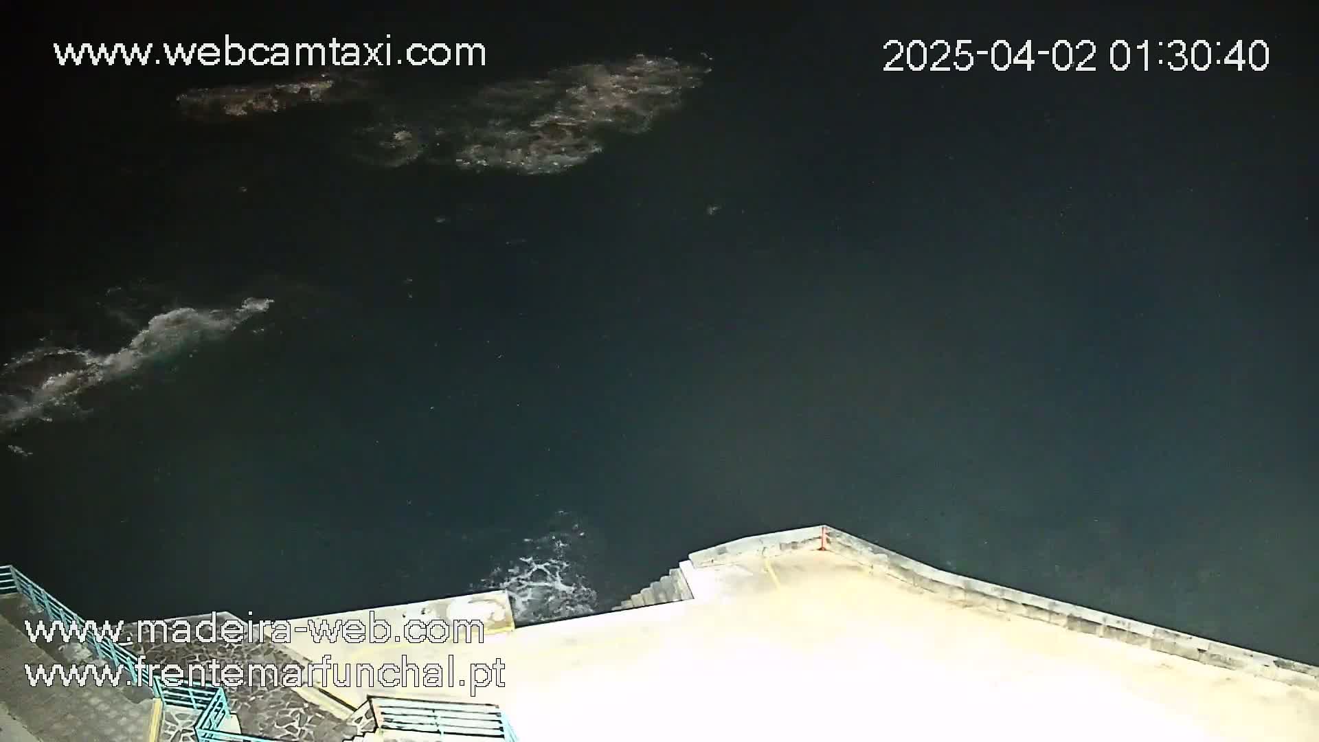 A nighttime, high-angle view shows dark water with small waves lapping against a light-colored concrete wall and steps.